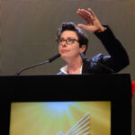 Image of Sue Perkins speaking at a lectern, raising one hand against a lit screen backdrop.