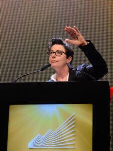 Image of Sue Perkins speaking at a lectern, raising one hand against a lit screen backdrop.