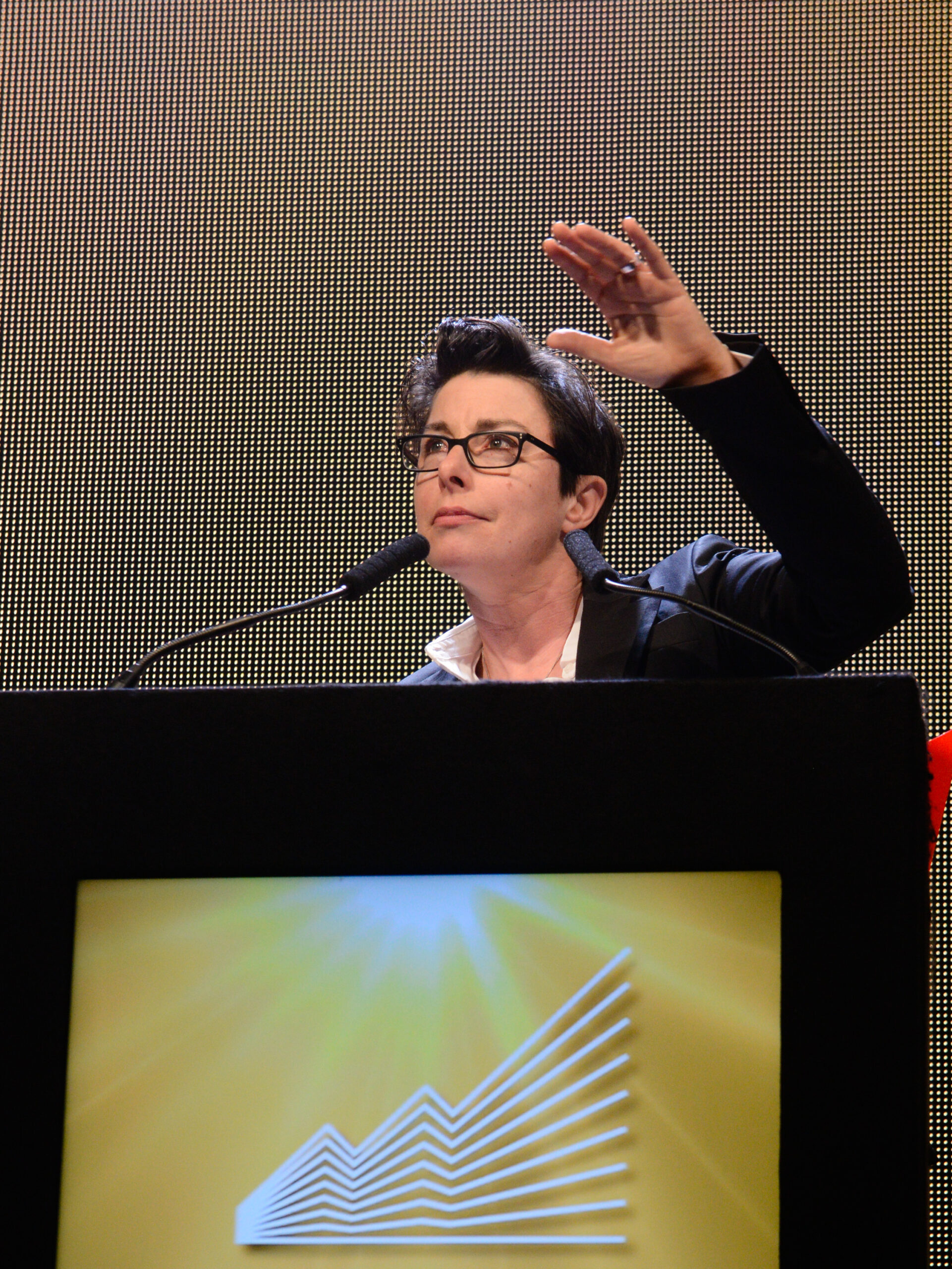 Sue Perkins SOMOs 2026 Image of Sue Perkins speaking at a lectern, raising one hand against a lit screen backdrop.